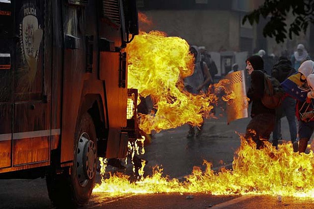 An anti-government protester stands with a shield near flames from molotov cocktails thrown at a water cannon by anti-government protesters during riots in Caracas